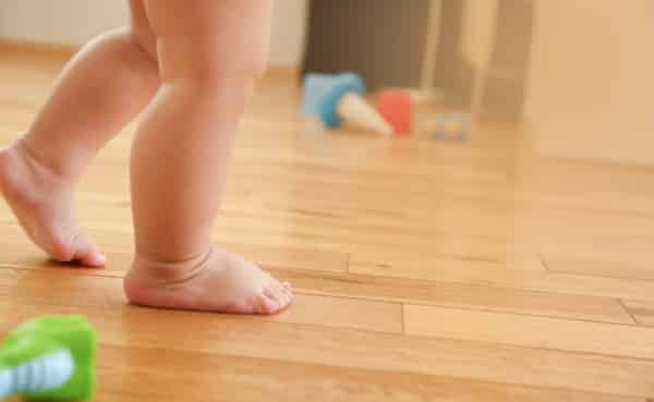 A toddler's bare feet taking baby steps on a wooden floor, with scattered toys in the background.
