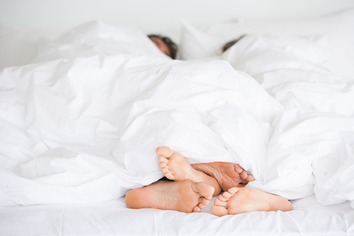 Two people's feet under a white blanket in bed, hinting at their relationship and sleeping habits.