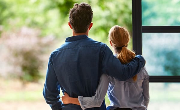 A man and a woman stand together by a window, facing outside. The man has his arm around the woman, embodying essential relationship tips for a stronger bond. Both dressed casually, they look out at the greenery visible through the window.
