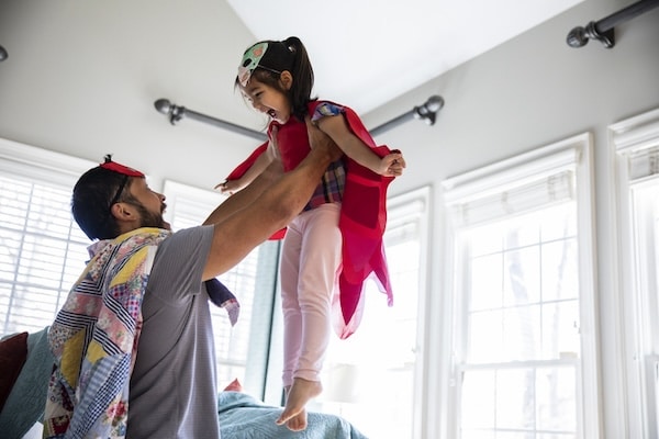 A dad is lifting a young child in the air. Both are wearing capes and masks, playing in a room with large windows. He embraces his essential role, making kids awesome through imaginative play.