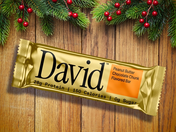 A gold-wrapped protein bar labeled "David" rests on a wooden surface with fir branches and red berries above. The peanut butter chocolate chunk flavor makes it a perfect stocking stuffer for men this holiday season.