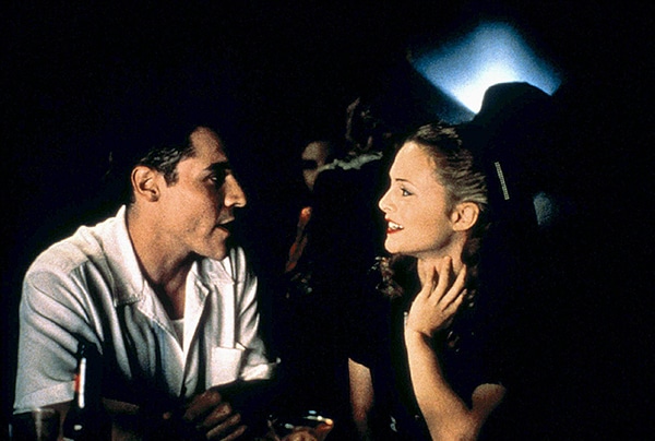 A man in a white shirt talks to a woman in dark clothing at a dimly lit bar; both appear engaged, exchanging classic 1995 flirting tips as they navigate how to flirt in the nostalgic nightlife setting.