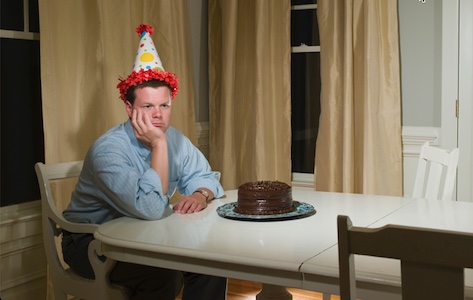 A man with a birthday cake on his head sits at a table.