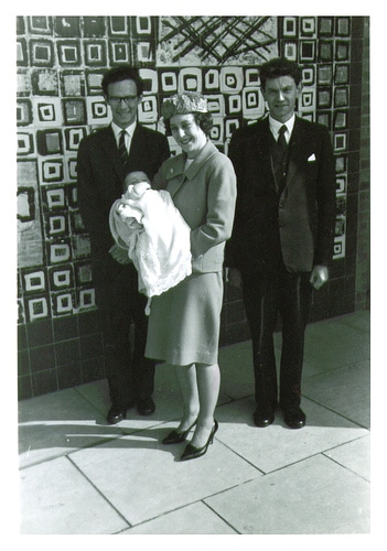 Three great individuals standing in front of a tiled wall with a baby in their arms.