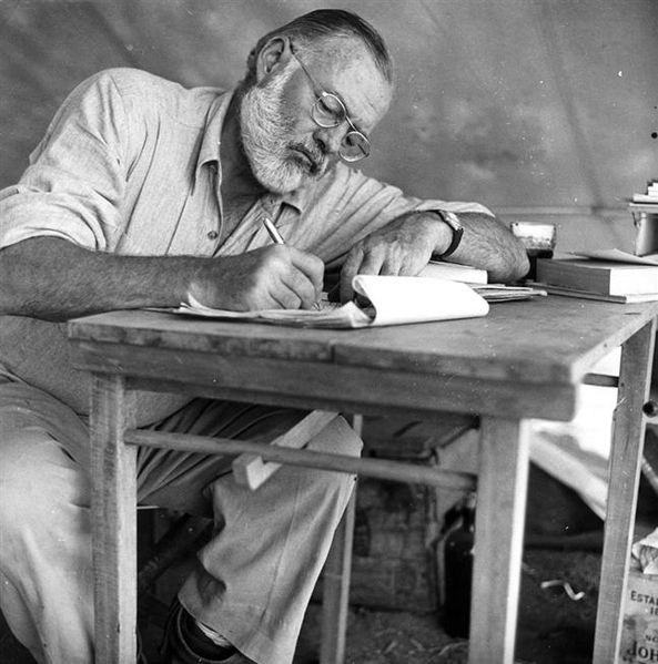 Ernest Hemingway, a man sitting at a table writing a book with motivational posters in the background.