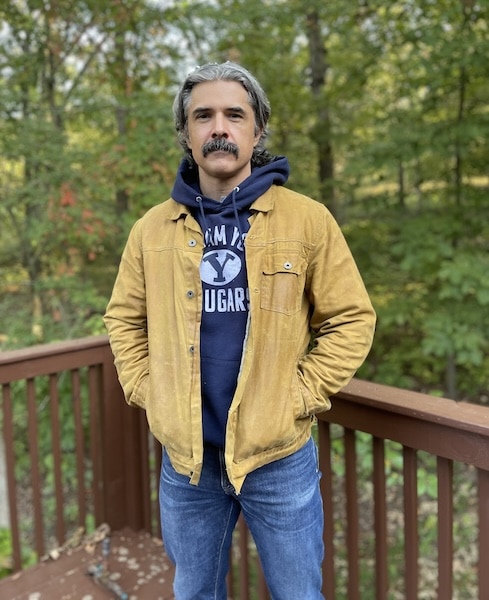 A man with gray hair and a mustache stands on a wooden deck, wearing a yellow fall jacket over a navy hoodie and blue jeans, with trees in the background.