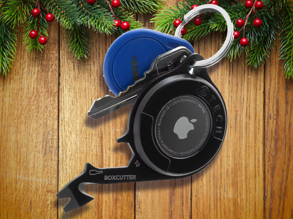 A keychain with keys, a blue fob, a black Apple AirTag, and a multi-tool rests on wooden boards with pine and red berries in the background—perfect stocking stuffers for men this holiday season.