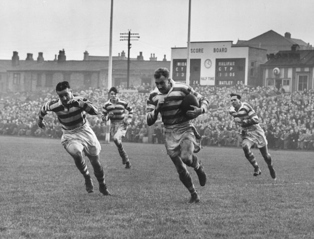 Four rugby players in striped Rugby shirts run across a grassy field during a match, with a scoreboard and large crowd of spectators in the background—a classic scene that highlights timeless men's fashion.