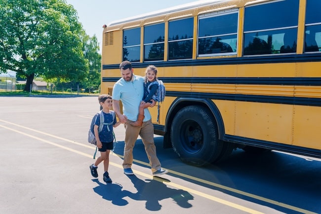 An adult dad walks with two children wearing backpacks toward a yellow school bus parked in a lot on a sunny, back-to-school day.