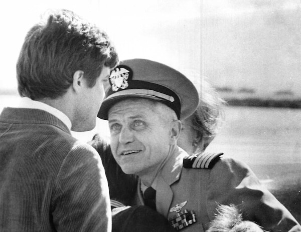 A man in a military uniform and cap, embodying resilience, speaks to another man in a jacket outdoors, with a blurred background.