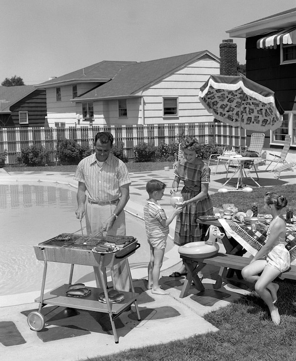 A family of four enjoys a backyard barbecue by a pool, capturing the essence of suburban life; the father showcases suburban manhood as he grills, while the mother and children stand nearby with patio furniture and an umbrella in the background.