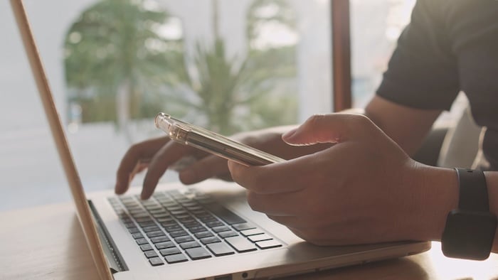 A person demonstrates task switching by using a smartphone in one hand while typing on a laptop with the other, seated at a desk near a window.
