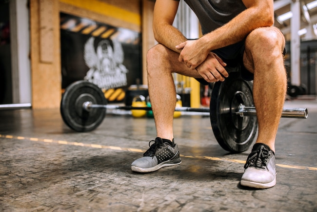 A person resting with hands on knees in a gym, with a barbell on the floor nearby, as if they don't feel like working out.