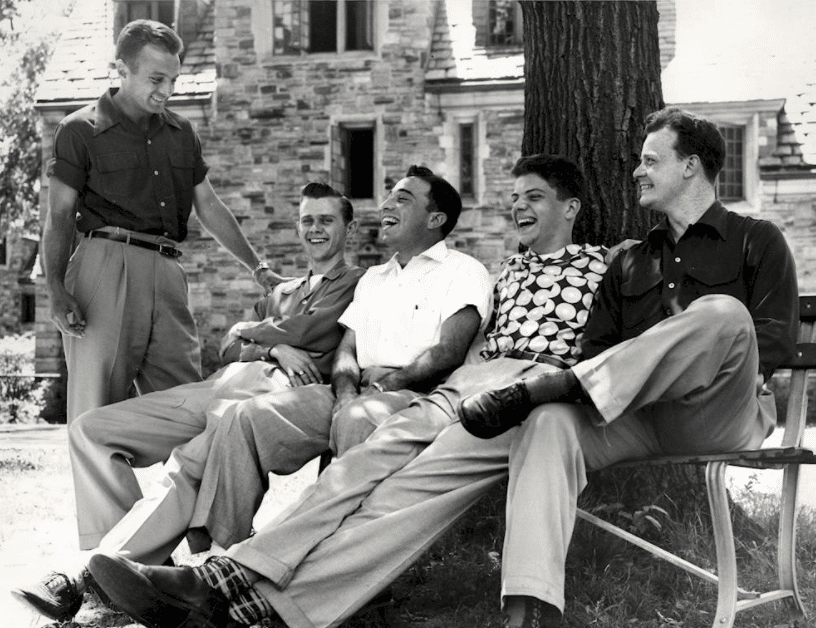 Several casual men are sitting on a bench in front of a building.